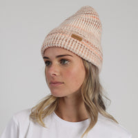 Woman wearing a beige knit beanie with a brand logo on a light gray background